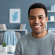 A smiling person, late 20s to early 30s, looking directly at the camera. The background is softly blurred with slate blue and
