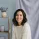 A happy, diverse woman in her late 30s smiles warmly, looking at the viewer. The background is softly blurred with sky blue a